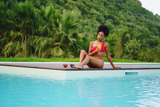 African american woman drinking smoothie and eating some fruit by the swimming pool - healthy lifestyle concept