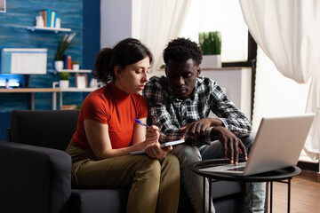 Focused caucasian woman writing on notebook while pensive african american man helping her. Diverse couple checking on banking bills and financial situation while discussing in living room at home.