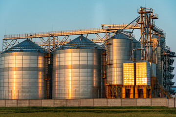 Obraz premium A large modern plant near a wheat field for the storage and processing of grain crops. view of the silver silos illuminated by the light of the setting sun against the blue sky. harvest season