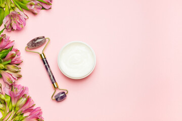 Cosmetic cream jar, face roller and flowers pink background. Skincare, natural organic cosmetics concept. Top view, flat lay, copy space.