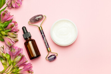 Obraz na płótnie Canvas Face roller with cosmetic serum and face cream on a pink background with flowers. Facial massage kit for lifting massage.