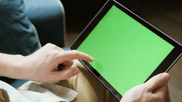 Young Guy Sits On A Chair And Watches A Video On A Tablet. Close-up Of A Man's Hand And Tablet With A Green Screen For Replacement. Turn On Play And Rewind Video On Your Tablet-PC.