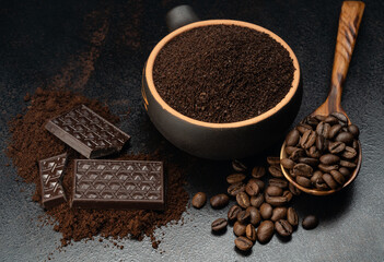 ground coffee in a cup with a spoonful of coffee beans and a bar of dark chocolate on a black background