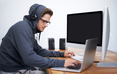 Engrossed in his work. A young hacker working on dual screens and a laptop wearing a hoodie.