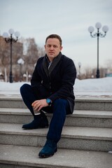 Obraz premium Young handsome man sits on concrete steps with coffee in a city park with lanterns, winter season, vertical photo