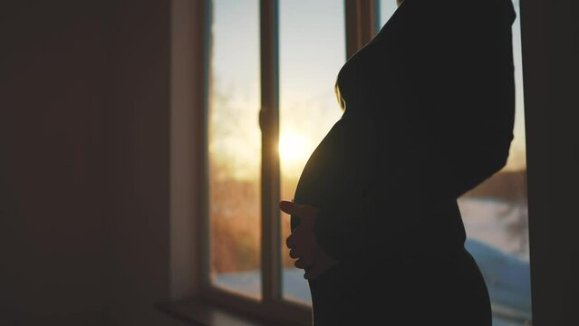 pregnant woman. motherhood a pregnancy concept. 40 year old pregnant woman stands in a dress by the window holding her stomach silhouette. silhouette of a girl in sunset by the window