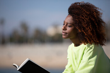 Side view of a young curly afro woman sitting on a breakwater holding a book while reading outdoors