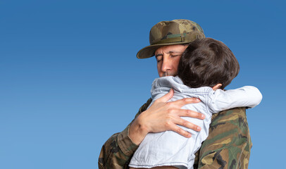 Military father hugs his son when they are reunited after a mission with a blue sky in the background and copy space.