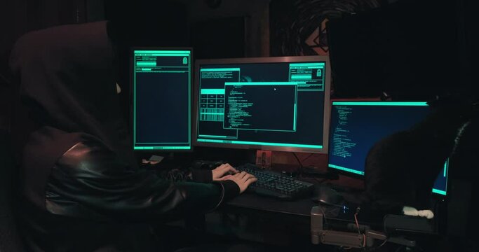 Hacker in a hood sits in front of computer screens and hacks databases. The concept of cyberterrorism, server hacking and cyberattacks. There is a black cat next to the man