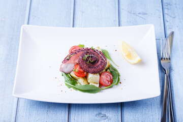 Warm salad with octopus, potatoes, cherry tomatoes, arugula, microgreens and lemon on a white plate. A traditional dish. Close-up, blue background.