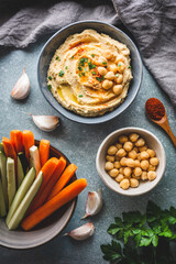 vista superior de un bol con hummus, un bol con verduras crudas, un bol con garbanzos, dientes de ajo y una cuchara de madera con pimentón, sobre fondo azulado