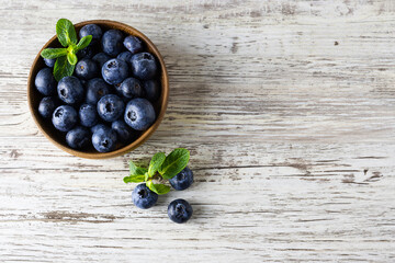Large and ripe blueberries in a wooden bowl on a light wooden table. Mint leaves on the table. Top view, space for text