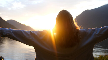 Woman raising hands on the edge of the mountain cliff on lake at sunset