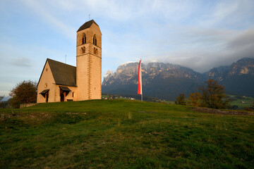 Die Kirche St. Peter am Bichl in Völs, liegt Malerisch auf einer Kuppe im Abendrot, vor dem Südtiroler Schlern, der von Wolken leicht umhüllt ist. - obrazy, fototapety, plakaty
