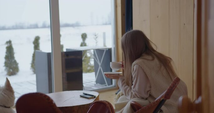 a fair-haired girl with long blond hair sitting in a blanket near the panoramic windows overlooking the winter forest drinks tea near the window with a white mug next to the dog white shepherd wolf 