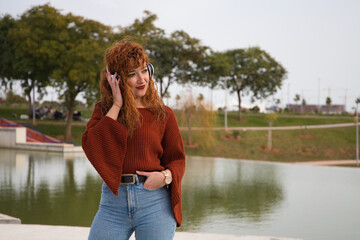 Young and beautiful red-haired woman listening to music with headphones in a park where there is a large lake. The woman is happy and dancing. Concept expressions and happiness, music and dancing.