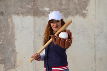 young and beautiful red-haired woman with baseball cap, jacket and glove with baseball bat resting...