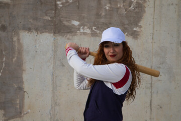 Obraz na płótnie Canvas young and beautiful redhead woman is happy with baseball cap, jacket and baseball bat in position to hit the ball. it is on grey cement background. Concept sport and recreations.