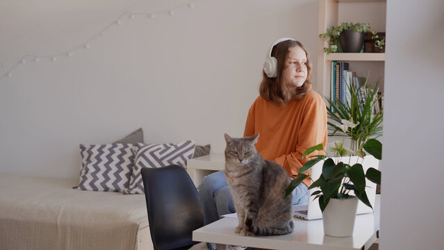 Teenage girl sitting on top of table in her room relaxing looking out the window, resting after studying at home online class talking to teacher via video call 