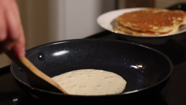 Hand flips pancake with wooden spatula in the kitchen with stack of pancakes in the background. Home cooking breakfast concept, dark moody shot.