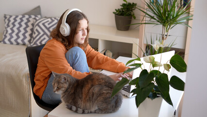 Teenage girl sitting on headphones with her cat nearby has school class online, interview with professor, video call with friends family at home.