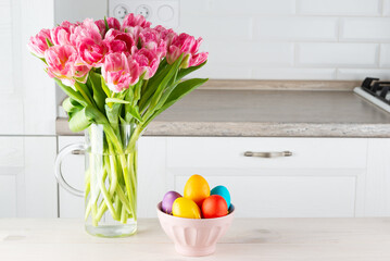 Easter colored eggs in a bowl with a bouquet of tulips on a wooden table.