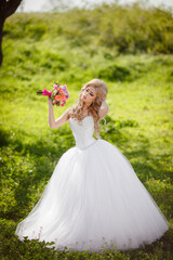 Obraz na płótnie Canvas A beautiful blonde bride with a cool make-up in a wedding dress holds a bridal bouquet against the backdrop of a beautiful, green nature. Husband and wife, happy together, just married, newlyweds.
