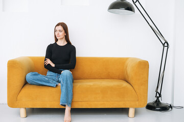 Young thinking woman girl with dark long hair in jeans sitting on yellow sofa in the minimalistic...