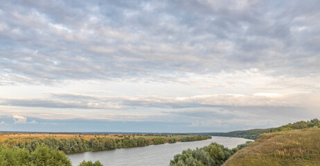 Scenic panoramic view. Green hills, fields and meadows. Sunny summer landscape with a river. Dramatic sky over the horizon. Calm. Clouds over the river.