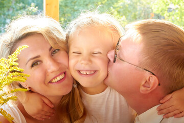 Portrait of Little blonde girl, mother and father on wooden terrace on nature or in the village and a green landscape in the background on a summer or autumn sunny day. Concept happy family
