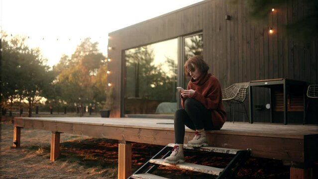 Young Cheerful woman resting and using phone on the wooden terrace near the modern house with panoramic windows near pine forest