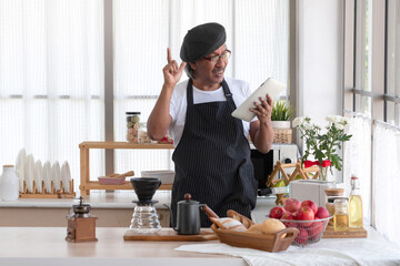 Senior man wearing black apron and hat doing a hobby with happy and relax using tablet for online social in modern kitchen