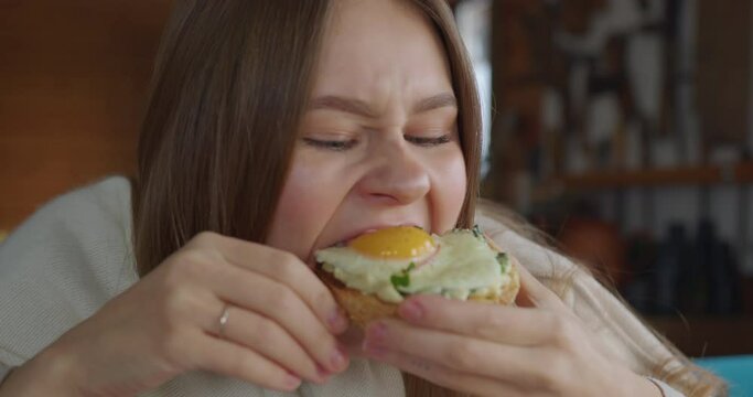 a girl with blond hair playfully look at the camera then eats a sandwich with a poached egg with great pleasure and chews very actively smiling. comically chews food like a hamster. close-up