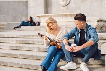 A music teacher teaches his young student to play the ukulele outdoors in the city.