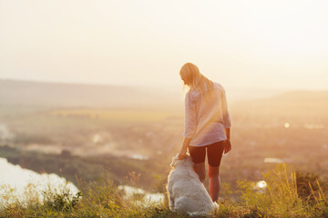 Blonde woman and her dog standing on hill and enjoying amazing view over river and mountains at sunset
