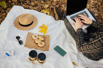 Hands man on laptop keyboard and coffee with donut on the picnic blanket in the autumn park. Easy freelance work life style.