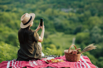 Young girl with hat making photo on smartphone at beautiful landscape while resting at summer picnic