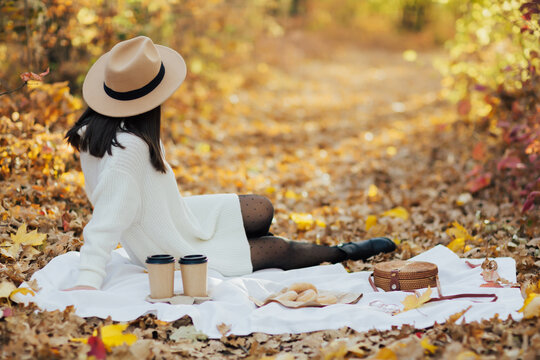 Young stylish woman sitting in the autumn park on the fall golden leaves and having picnic with sweet donuts and coffee 