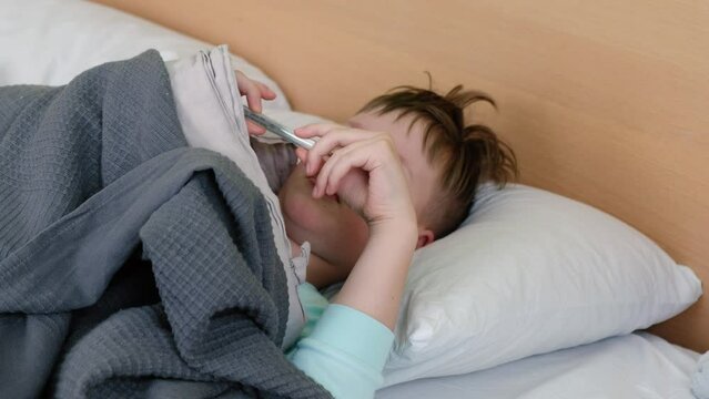 A sad boy is lying on the bed and measuring the temperature with a mercury thermometer, and he has a headache. The face is distorted with pain. High quality 4k footage