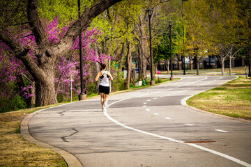 Woman in shorts with hair up running on curved paved running path through park in springtime with...