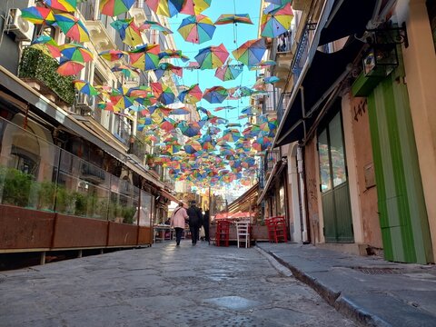 Stradina caratteristica in basolato del centro di Catania, con decorazioni con ombrelli colorati appesi tra i palazzi antichi, e persone che camminano