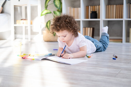 curly child girl drawing with colored markers lying on floor in living room at home