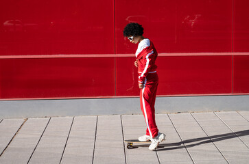 Young African American woman with short hair and sportswear with her skateboard over a red colored wall