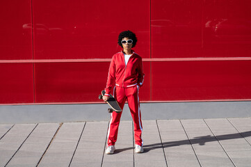 Young African American woman with short hair and sportswear with her skateboard over a red colored wall