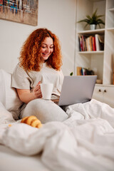 A happy redhead girl sits on her bed in the bedroom in the morning with a laptop in her lap and drinks coffee. A girl using the laptop for browsing the web.