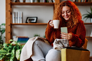 A cute ginger girl with curly hair is sitting in the chair at home in the morning, texting messages...