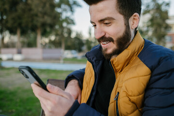Young man looking at the phone, handsome man smiling looking at his phone, man wearing coat and sitting outside in cold weather