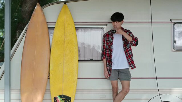 Young asian man standing with drinking hot coffee and surfboard at motor home