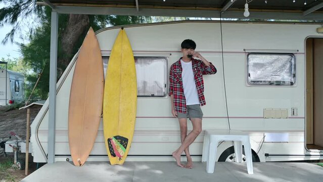 Young asian man standing with drinking hot coffee and surfboard at motor home