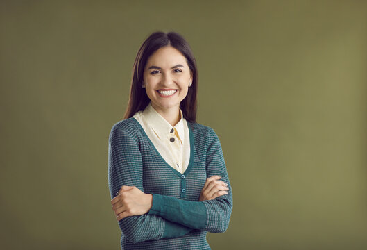 Studio portrait of happy woman with pretty face and positive, charming smile. Cheerful, smiling good-looking young lady in blouse and jumper standing with her arms crossed isolated on green background